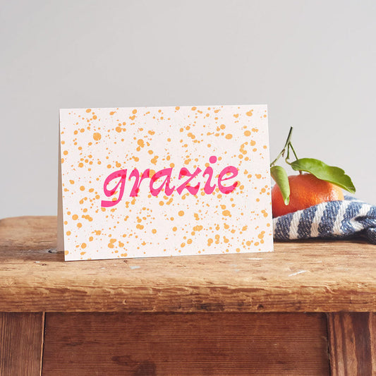 Card with 'grazie' on a wooden surface with a fruit and fabric background